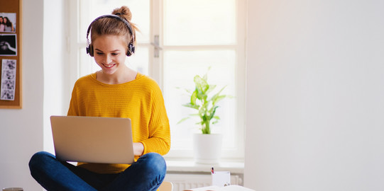 A woman sits cross-legged on a desk with a laptop on her lap.