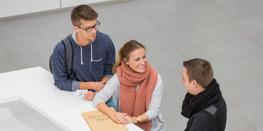 Drei Personen stehen neben einenander an einer Theke. Die Person in der Mitte lacht die Person rechts an und hat ihre Hände auf einen großen Umschlag liegen. Die Person links hat einen kleinen Zettel mit einer Wartenummer in der Hand.
