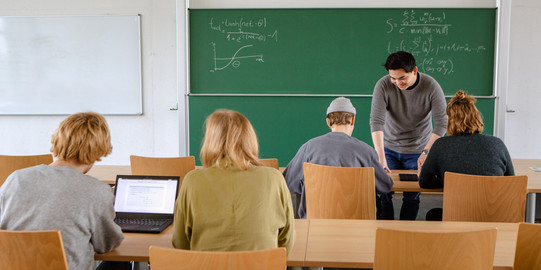 Vier Studierende sitzen im Seminarraum zur Tafel gewandt. Der Dozierende erklärt etwas.
