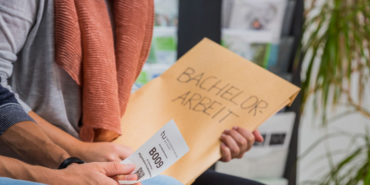 Zwei Personen sitzen nebeneinander. Man sieht einen Ausschnitt von Knie bis Brust. Die hintere Person hat einen großen Umschlag mit der Aufschrift „Bachelor-Arbeit“ in der Hand, die vordere Person hat einen kleinen weißen Zettel mit einer Wartenummer, einem Code und dem TU-Logo in der Hand.