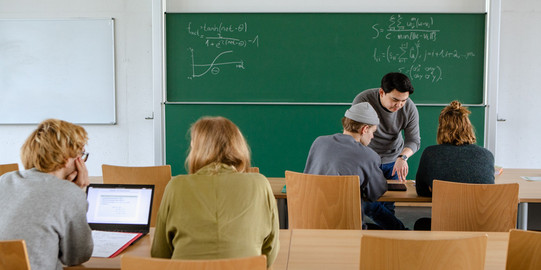 Vier Studierende im Seminarraum sitzen und arbeiten zusammen mit dem Dozierenden im intergrund.