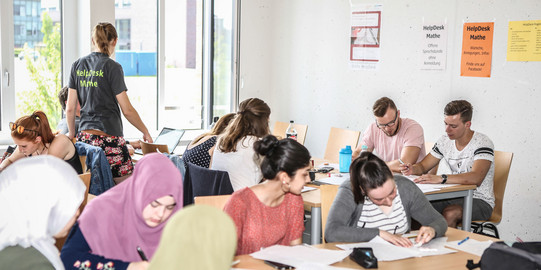 Mehrere Personen sitzen an Tischen und haben Unterlagen vor sich. Eine Person steht und trägt ein T-Shirt mit der Aufschrift "HelpDesk-Mathe".